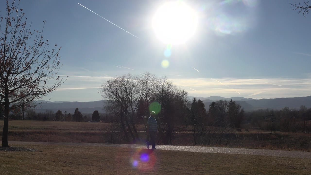 Older man walking on a paved path under bright sun beside open grass and bare trees.