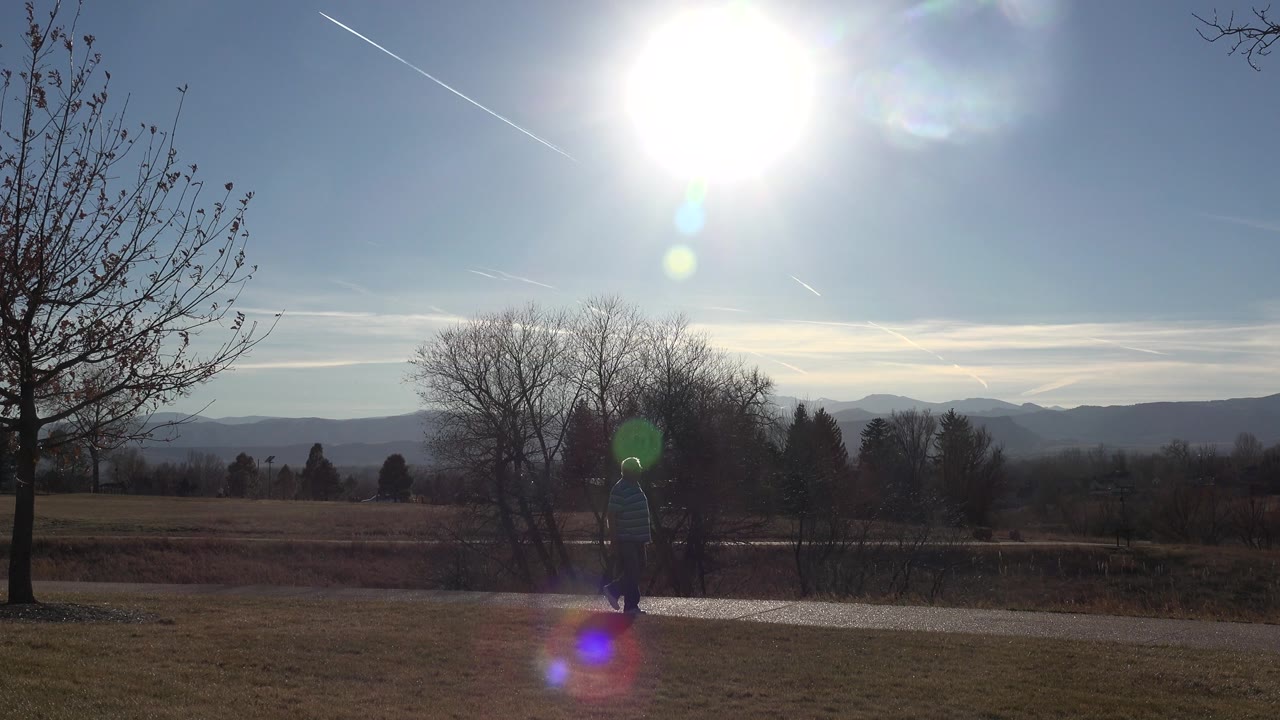 One second in, the older man remains small in the frame on the Arvada path, with the bright sun high overhead, green and purple lens flare, bare trees, and layered hills behind him.