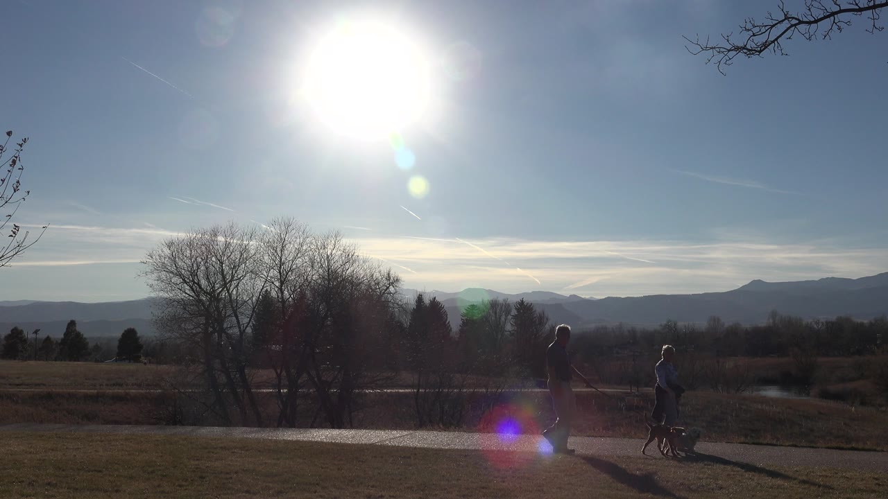 At the start of the Arvada, Colorado clip, two backlit older adults walk small dogs along a path with bare trees and distant mountains behind them.