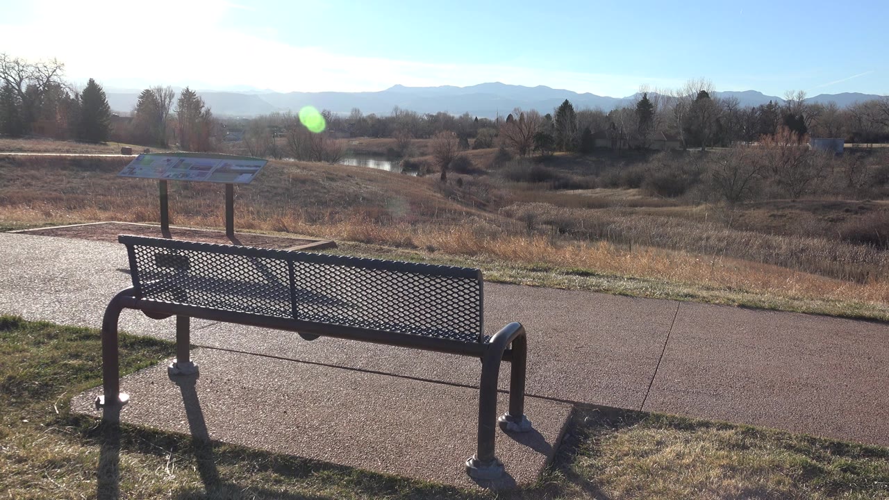 Black metal bench beside paved paths with dry grass and distant mountains.