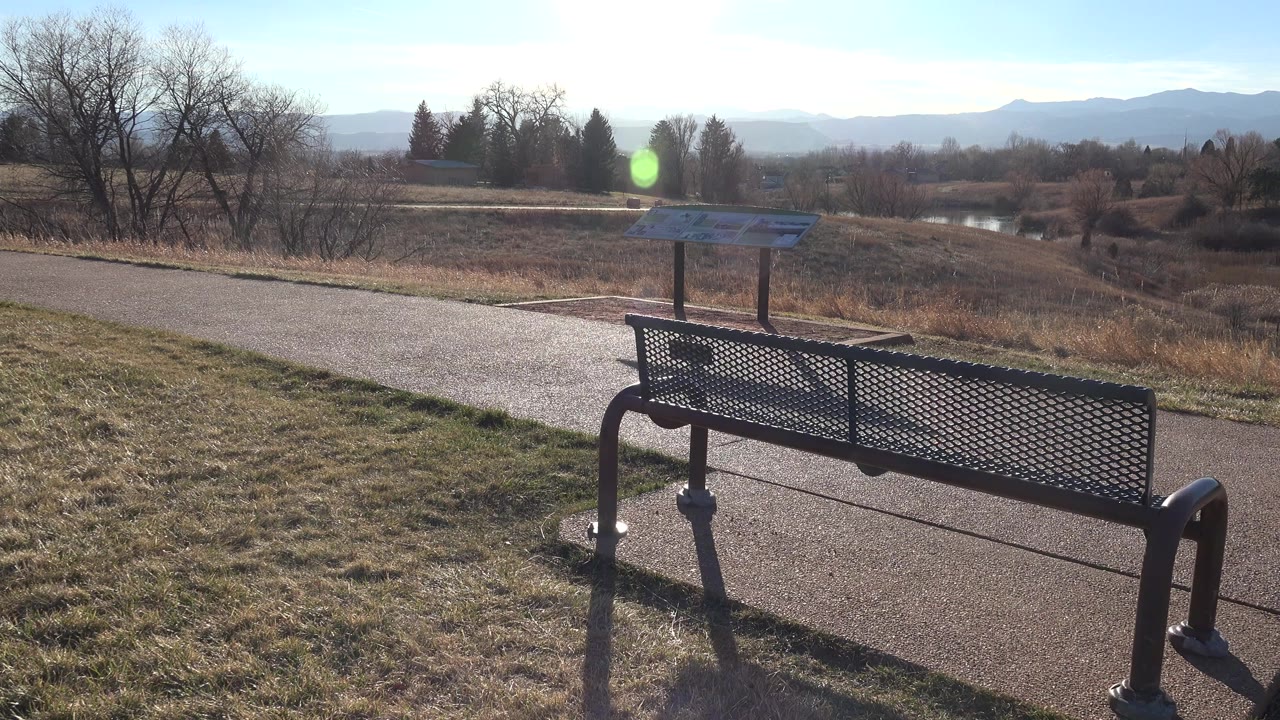 At 0:00 in Arvada, Colorado, a dark metal bench anchors the foreground beside a paved path, with an interpretive sign, dry field, leafless trees, distant water, and no visible people.