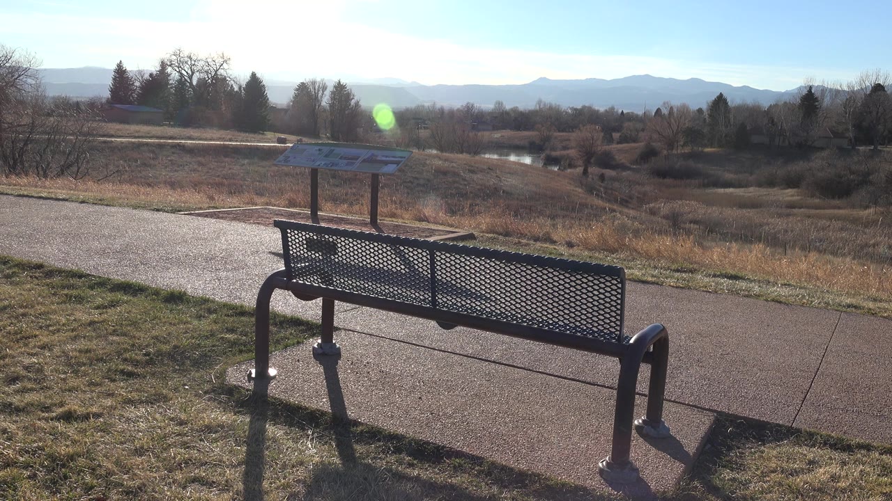 At 0:00 in Arvada, Colorado, a black metal bench fills the foreground beside a paved path, with an interpretive sign, dry reserve grass, bare trees, distant mountains, bright sun glare, and no visible people.