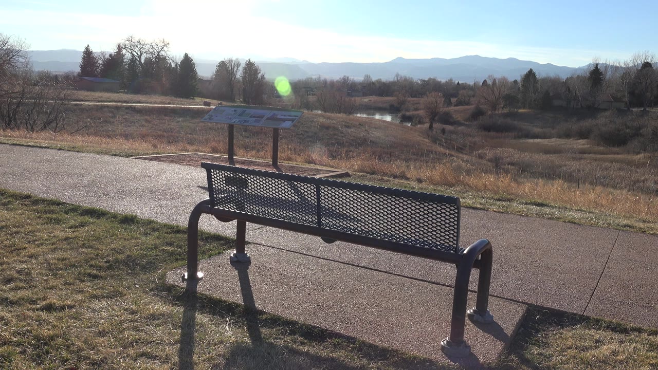 At 0:01 in Arvada, Colorado, the bench remains low in the foreground while the sign stands behind it near the path, with open brown grassland, tree lines, mountain silhouettes, a green lens flare, and no visible people.
