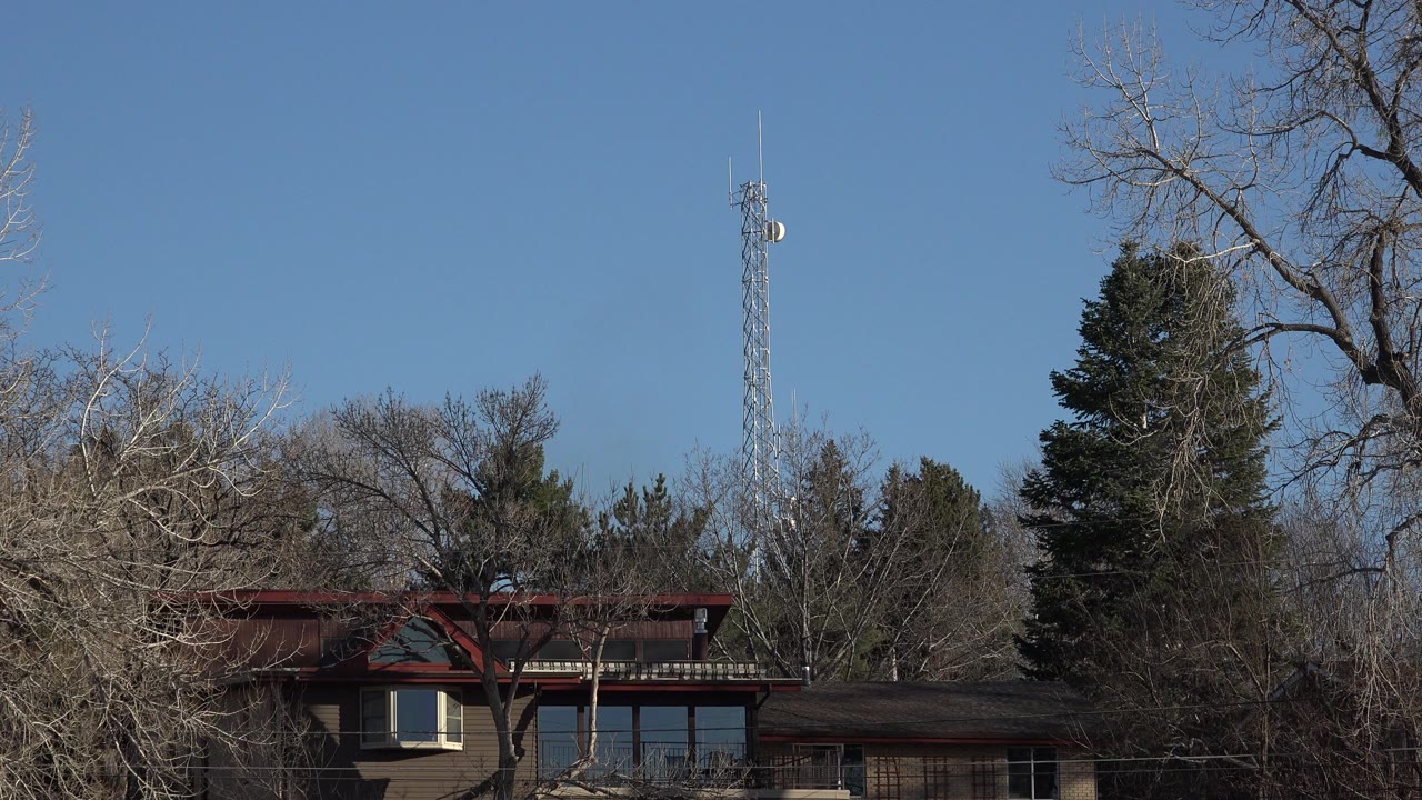 Second frame shows the Arvada residential facade more centered behind winter-bare branches, with dark evergreens and a slim tower against the blue sky and no visible people.