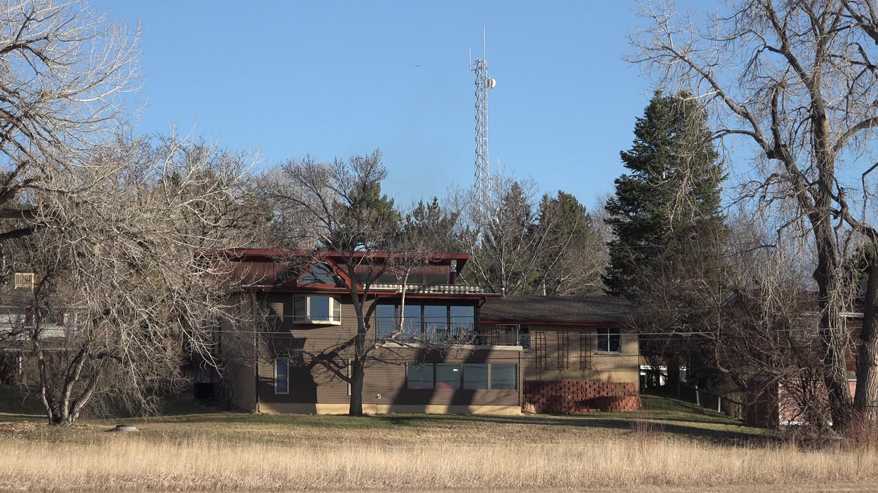 Tan house in Arvada, Colorado seen beyond a dry grassy foreground, bare trees, evergreens, and a tower in clear daylight, with no visible people.
