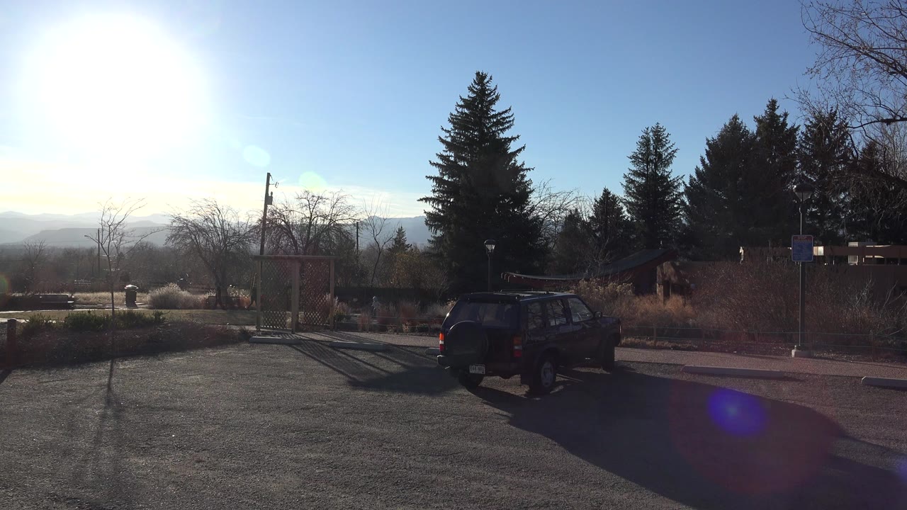 Bright sun over a park edge with a dark SUV, playground structure, trees, and distant hills.
