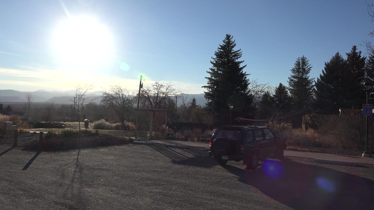 The red car remains parked on the right side of the Arvada, Colorado frame, with the sun high over the parking area and low hills visible beyond the trees.