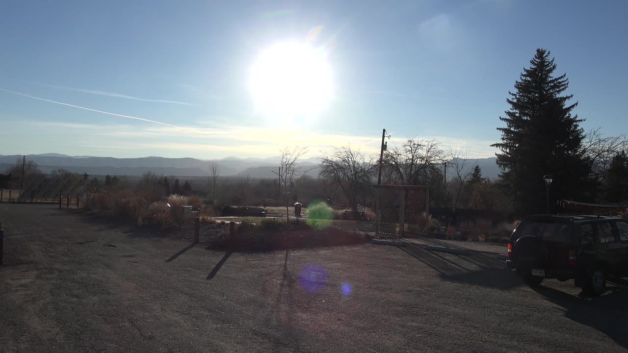 A bright Arvada, Colorado outdoor view shows the sun above low clouds, a dark vehicle near the right edge, driveway pavement, shrubs, and no visible people.