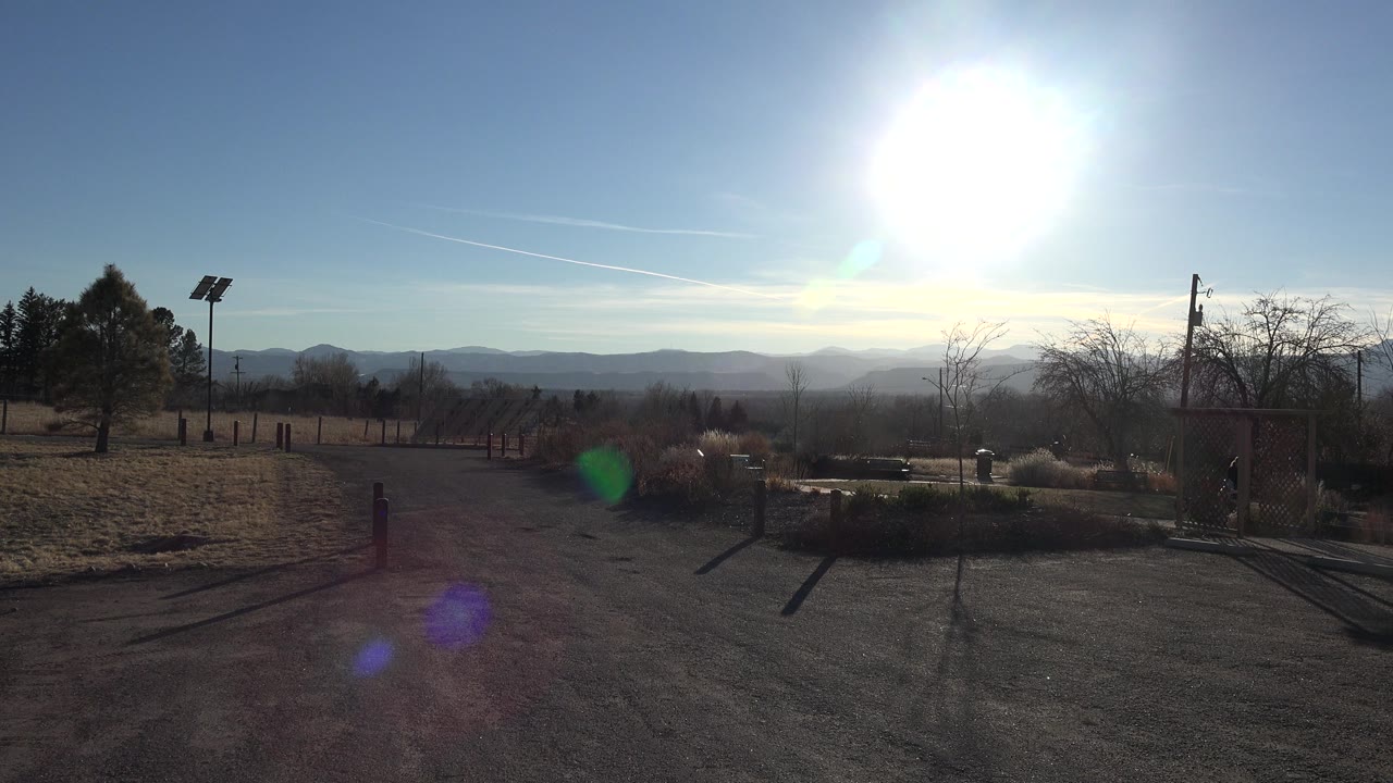 Opening frame in Arvada, Colorado showing a paved drive, frosty grass, fence posts, distant hills, bright blue sky, strong sun flare, and no visible people.