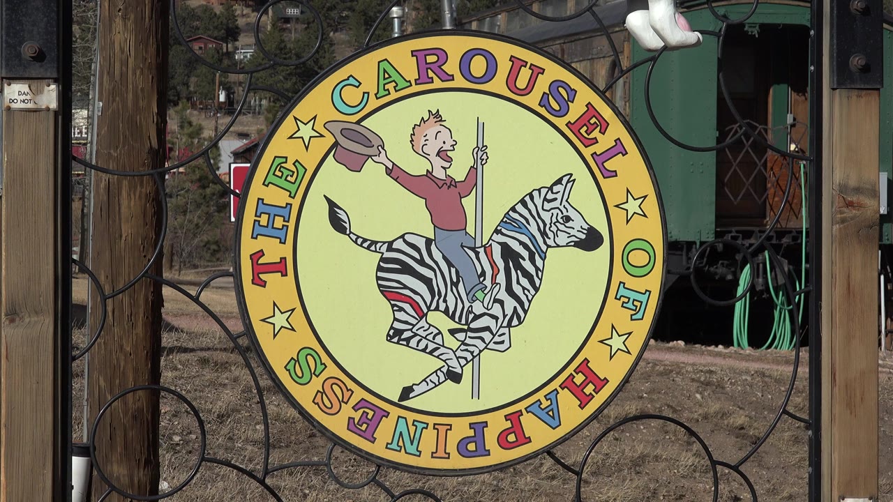 At 0:00, a yellow Carousel of Happiness sign with a child riding a zebra hangs on a black metal gate in Nederland, Colorado, with wooden posts and no visible people.