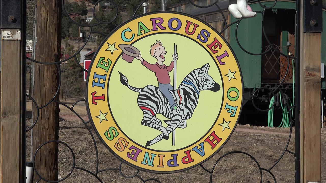 At 0:01, the circular Carousel of Happiness sign remains centered behind black gate loops, with the zebra illustration, wood posts, and utility area visible behind it in Nederland, Colorado.