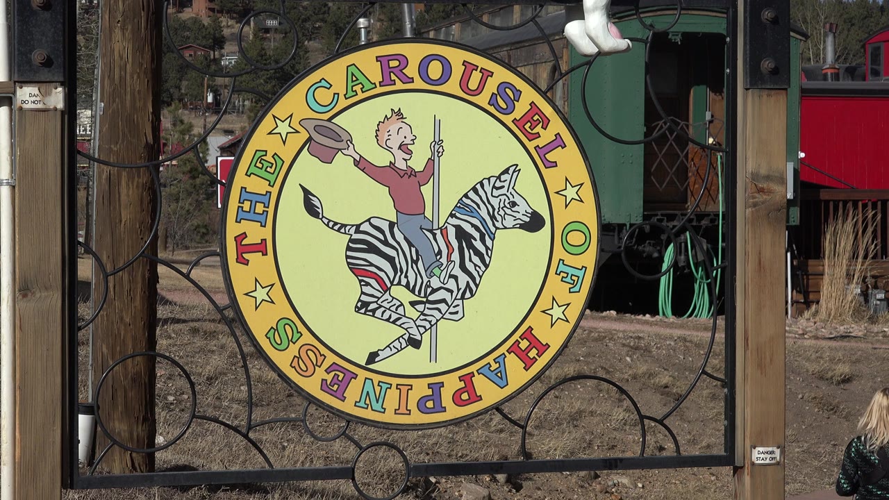 The Carousel of Happiness sign remains centered in Nederland, Colorado, with a partial blonde person visible at the lower right edge near the walkway and red and green buildings behind it.