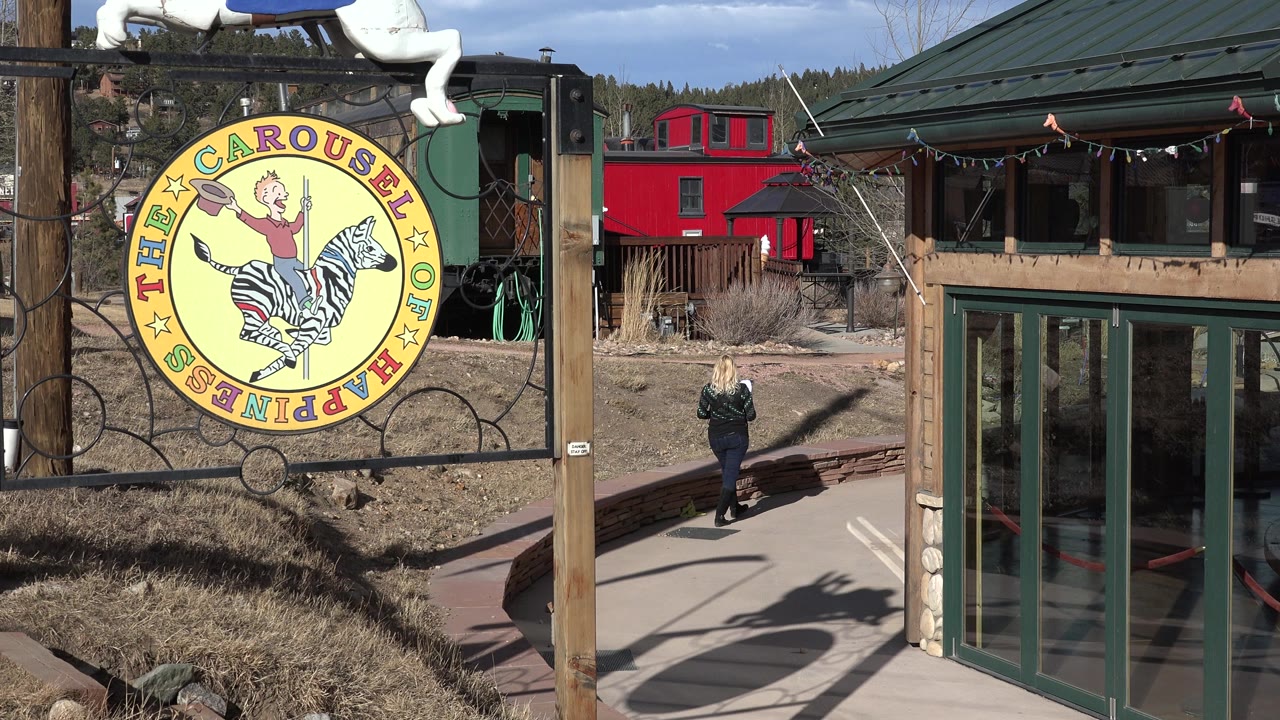 Nederland, Colorado frame showing a large yellow carousel sign, glass-fronted building, dry grasses, and a woman holding a child's hand near the center path.