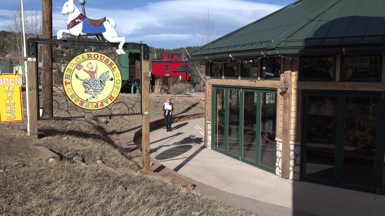 At 0:01 in Nederland, Colorado, the carousel-themed sign remains at left while the person stands farther along the sunlit walkway near the green-framed glass doors.