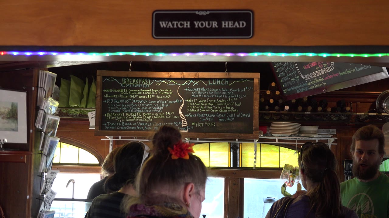 Patrons stand below a chalkboard menu inside a wood-paneled cafe.