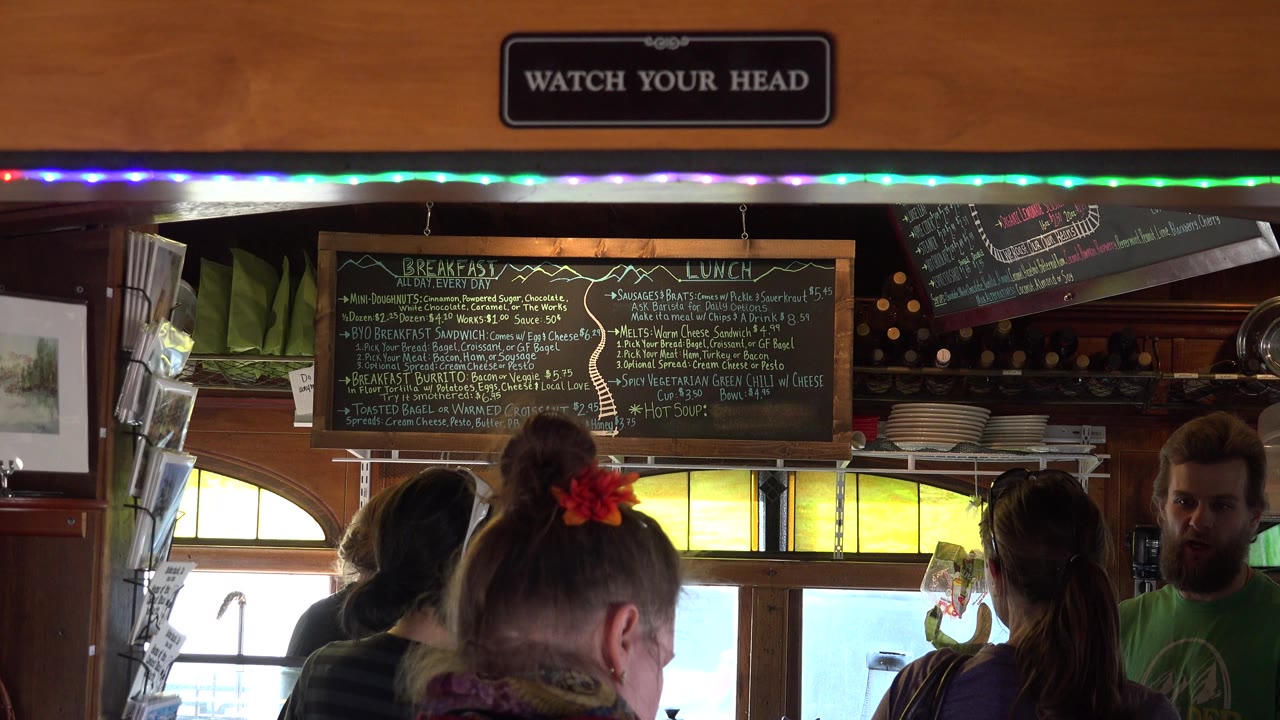 In Nederland, Colorado, several people stand with their backs to the camera while a bearded man at right is visible near the counter and chalkboard menu.