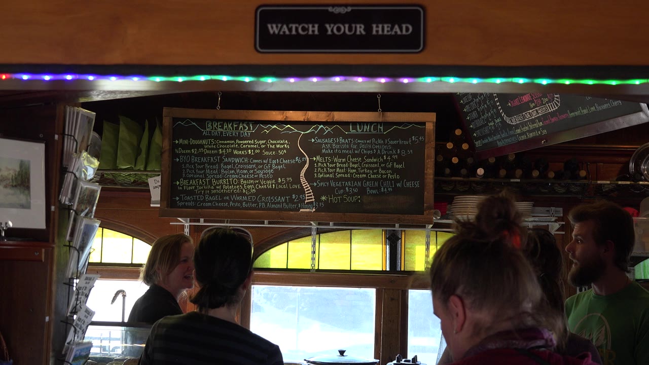 Customers at a restaurant counter beneath a chalkboard menu and watch your head sign.