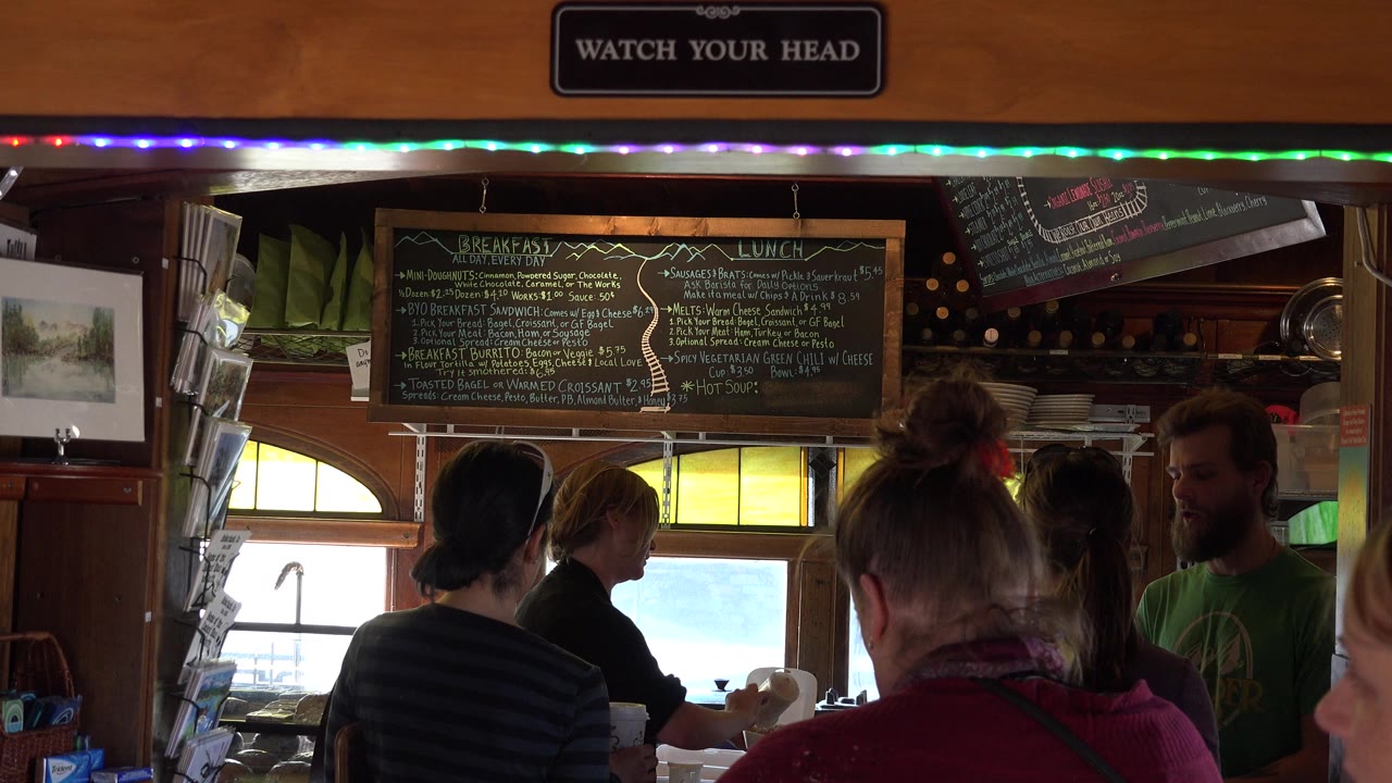 In Nederland, Colorado, several people stand with their backs to the camera inside a wood-paneled public interior beneath a chalkboard breakfast and lunch menu.
