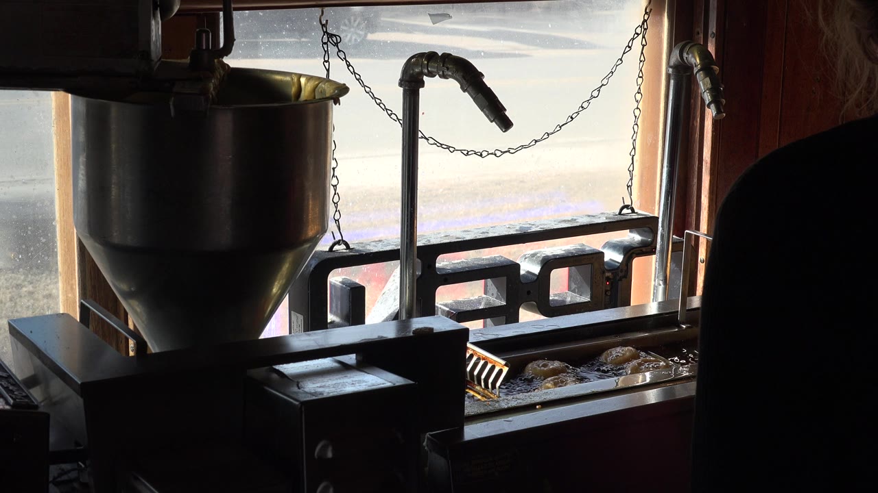 A metal hopper, angled pipe, chain, and fryer tray are silhouetted against the window in the Nederland, Colorado food prep interior, with a dark partial figure at left.