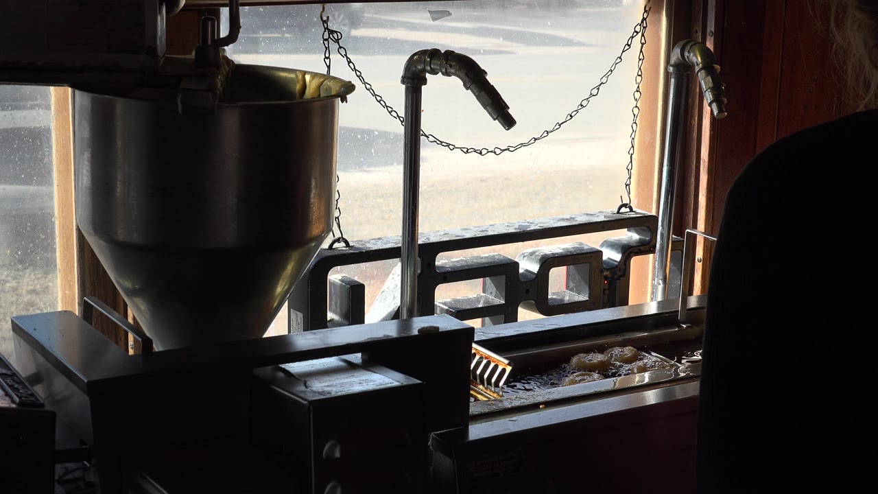 The fryer setup remains silhouetted near the window in Nederland, Colorado, while round fried items sit along the lower tray and the partial figure stays unidentifiable.