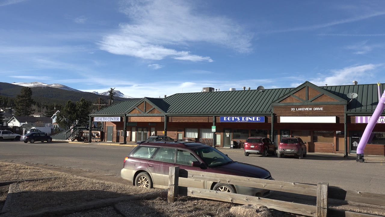 Shopping strip parking lot with cars, storefronts, mountains, and blue sky.