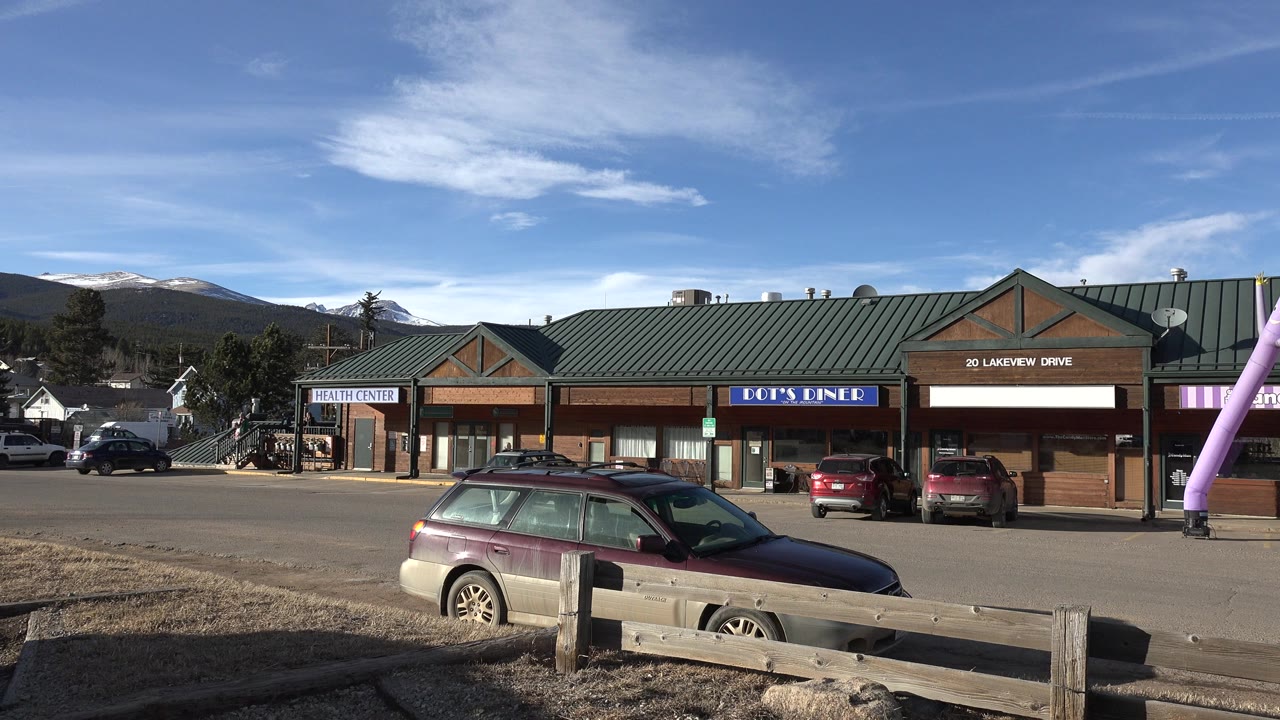 Nederland, Colorado storefront row with Dot's Diner signage, parked cars, a foreground wood rail, and the pink inflatable tube figure partly visible at the right edge.