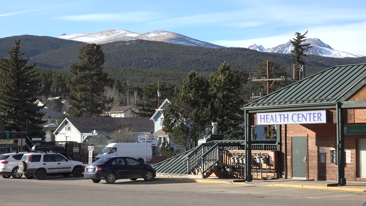 Health center building with parked cars and snowy mountains behind town trees.