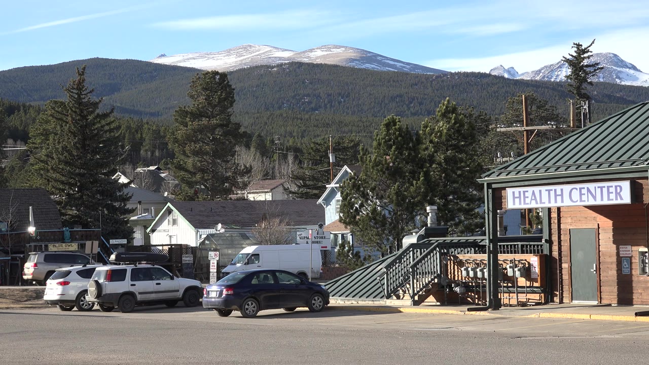 Nederland, Colorado street frame showing a white SUV, dark car, brick Health Center facade, trees, rooftops, and a snowy mountain ridge under bright daylight, with no visible people.