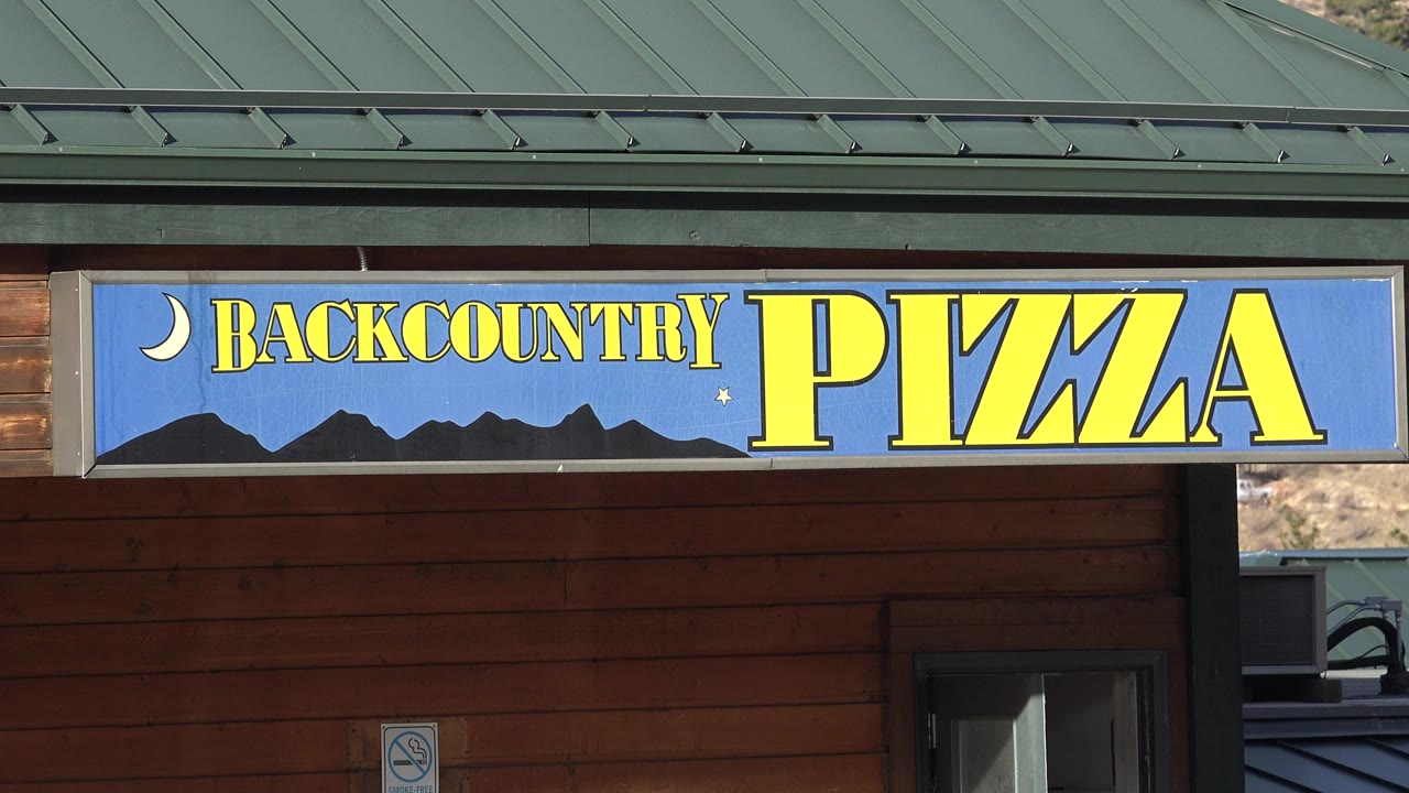 Backcountry Pizza sign on a wood building under a green metal roof.