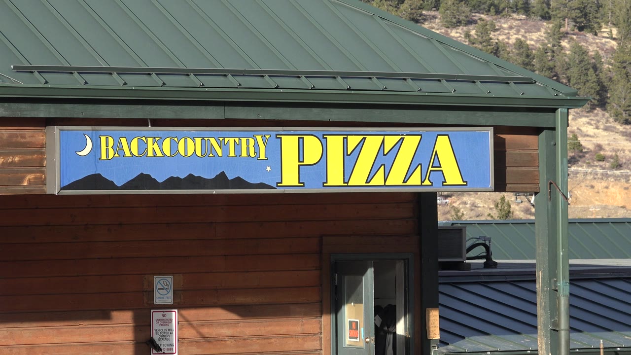 Slightly wider view of the Backcountry Pizza facade in Nederland, Colorado, showing more of the roof, entry area, hillside, and no visible people.