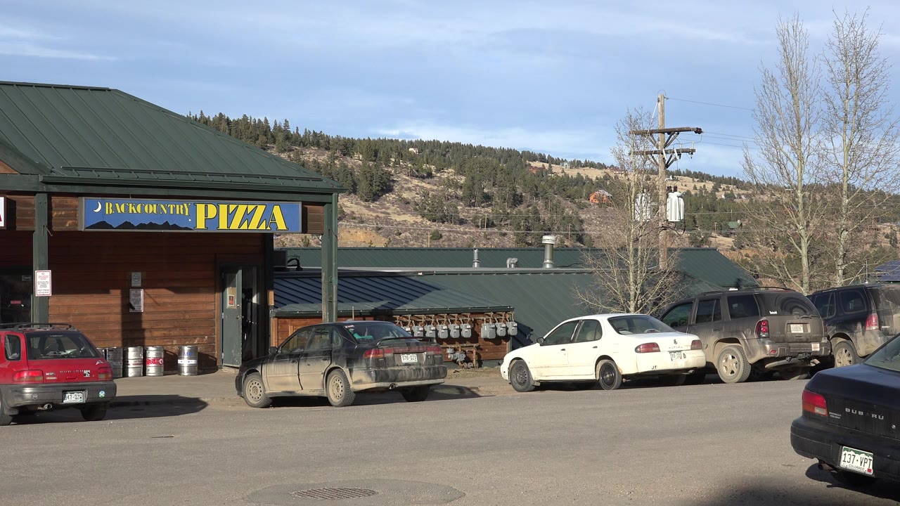 Small town parking lot outside shops with parked cars and hills behind.