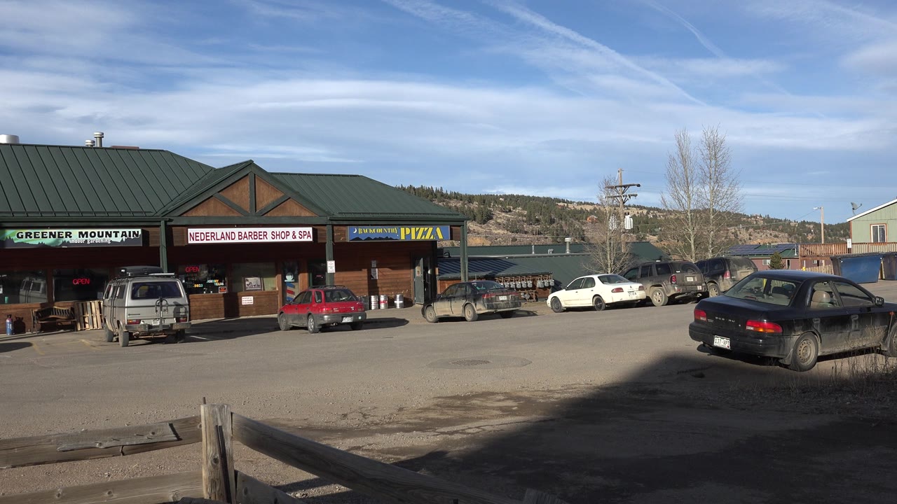 Strip mall storefronts with parked cars under a blue sky.