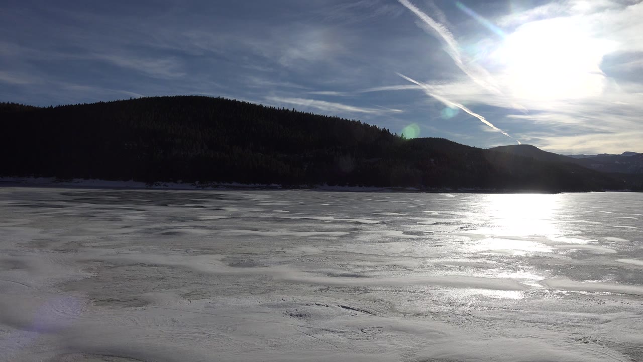 Opening frame of an icy lake in Nederland, Colorado with bright sun glare, dark wooded hills on the horizon, wispy clouds above, and no visible people.