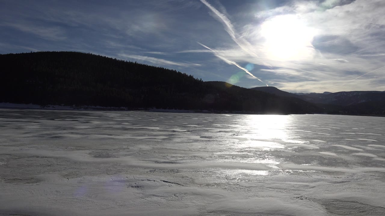Wide view of a frozen lake in Nederland, Colorado with bright sun reflecting off the ice, dark mountains on the horizon, streaked clouds, and no visible people.