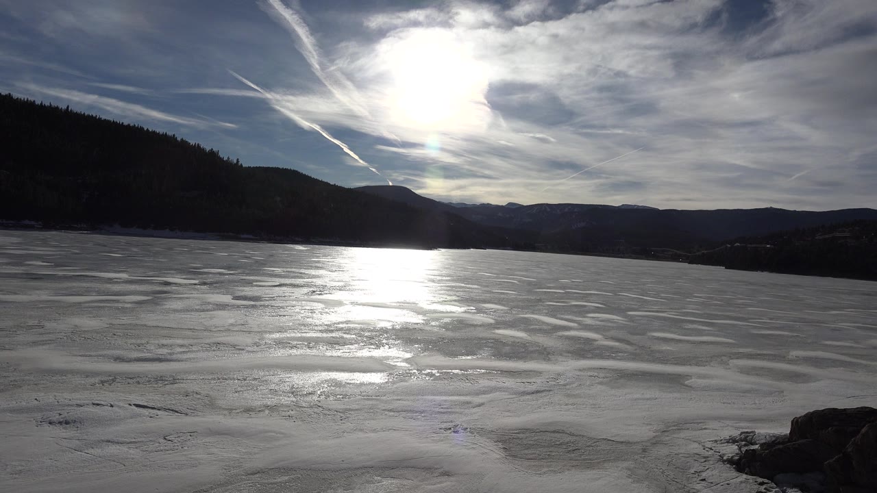 A frozen lake in Nederland, Colorado fills the foreground under a bright sun, with dark mountain slopes on the left and no visible people.
