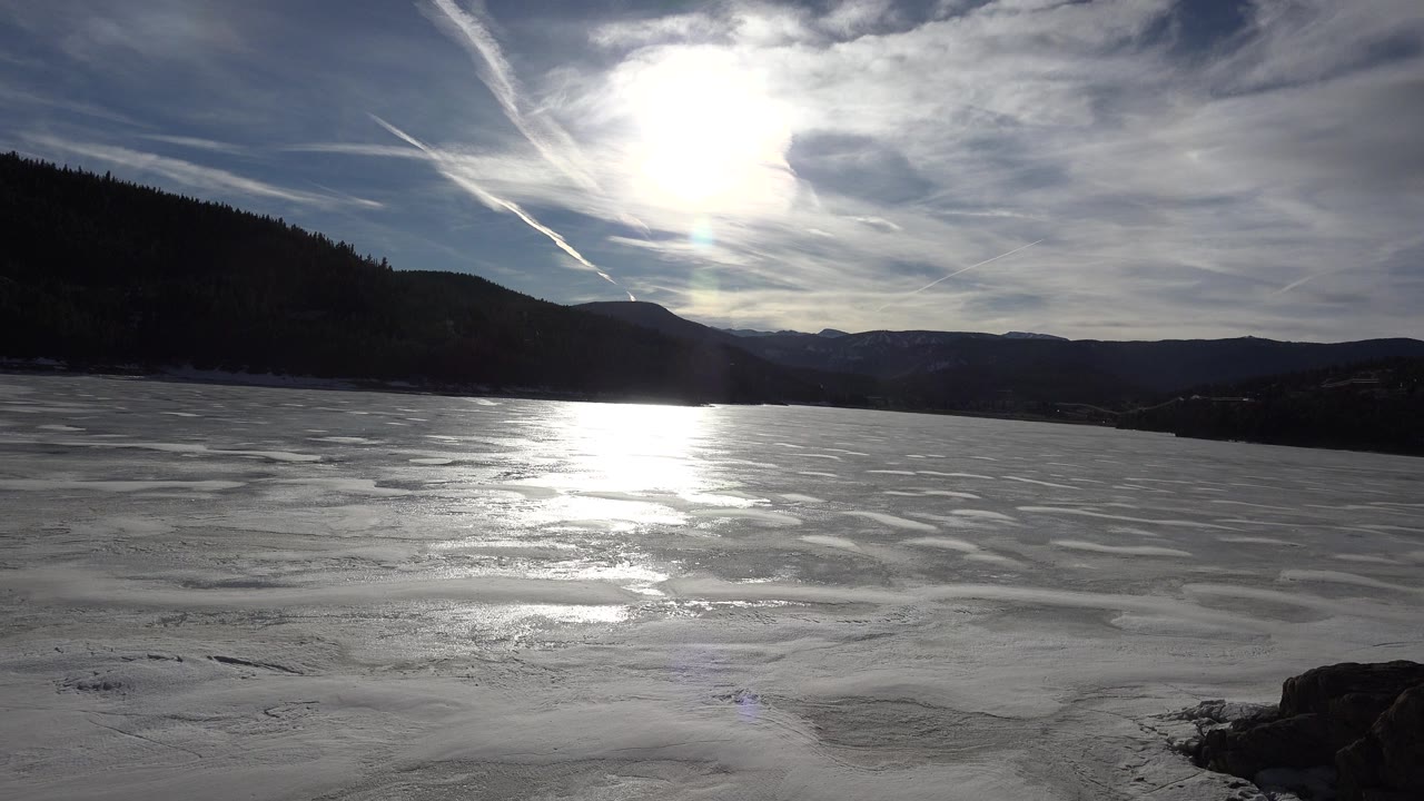 Bright sunlight reflects across rippled ice on a frozen Nederland, Colorado lake, with layered ridgelines beyond and no visible people.