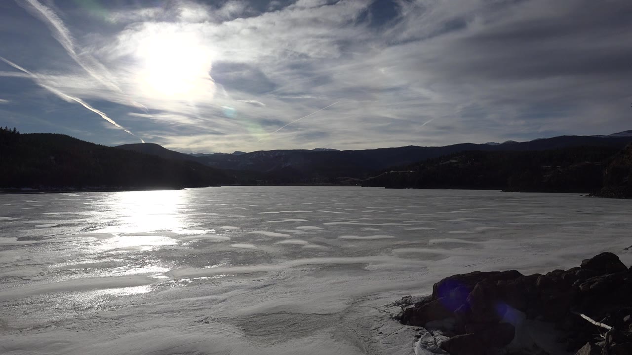 Nederland, Colorado landscape frame with hard sun glare above a pale rippled water or ice surface, dark hills on the horizon, rocks at lower right, and no visible people.