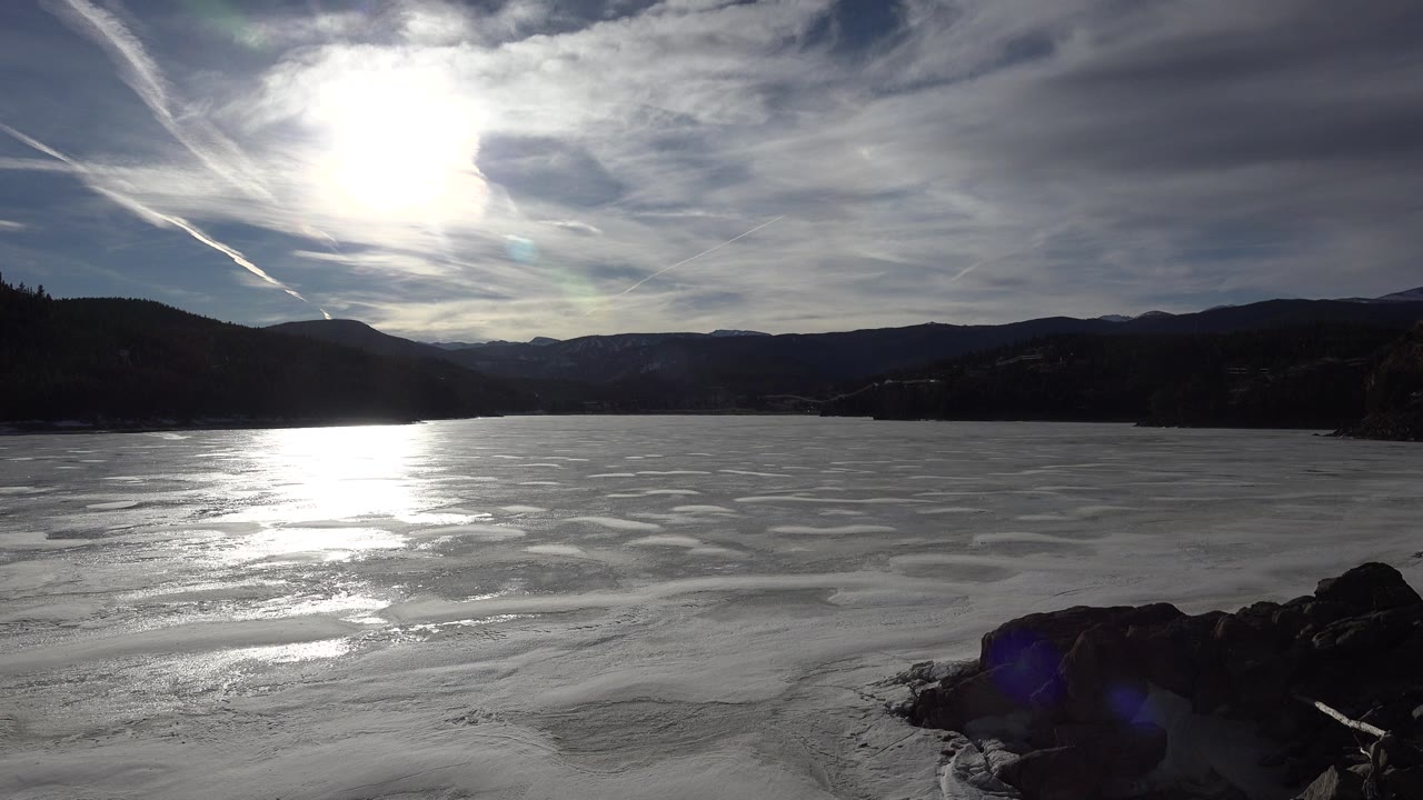 Wide view of frozen water in Nederland, Colorado with bright sun reflection on the ice, dark rocks at the lower right, distant mountain ridges, and no visible people.