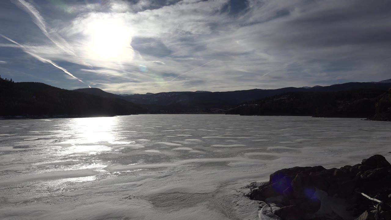 Frozen water in Nederland, Colorado reflects a bright patch of sunlight while dark wooded hills and low mountains stretch across the background with no visible people.