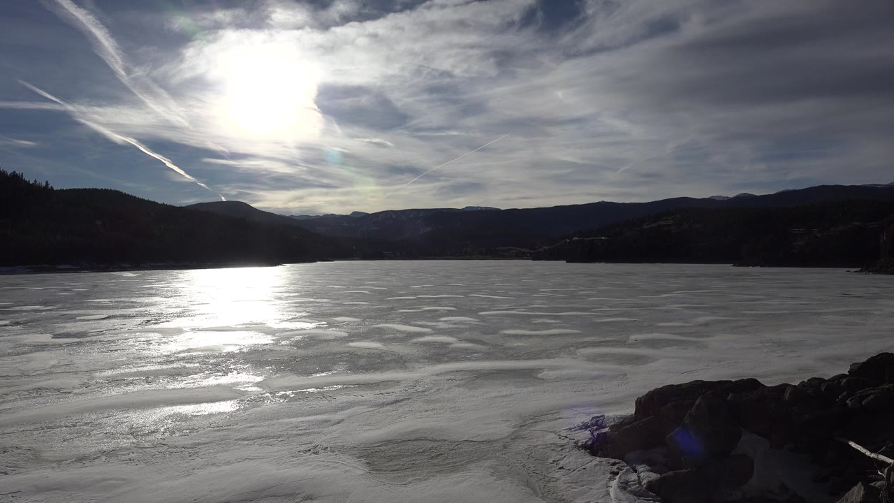 Bright sun over frozen water with a dark mountain ridge and rocks at right.