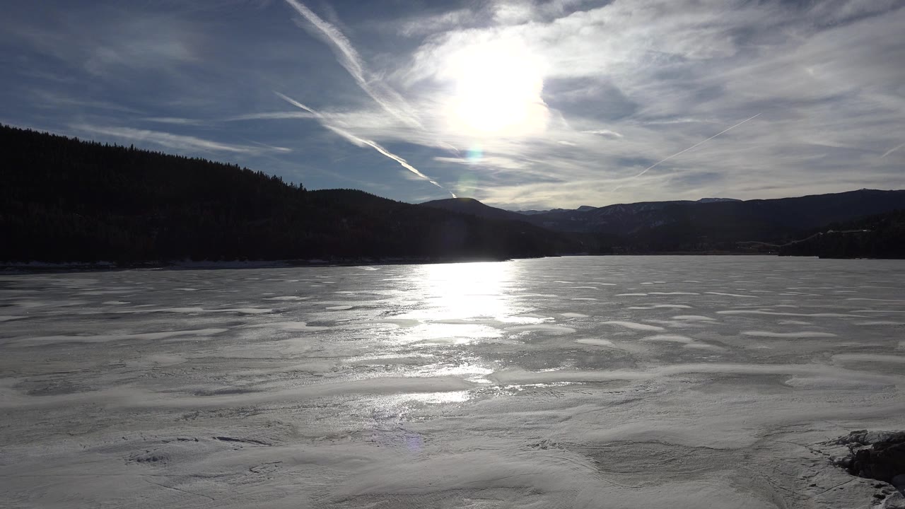 Frozen lake in Nederland, Colorado with bright sun reflecting on the ice, dark wooded hills on both sides, streaked clouds overhead, and no visible people.