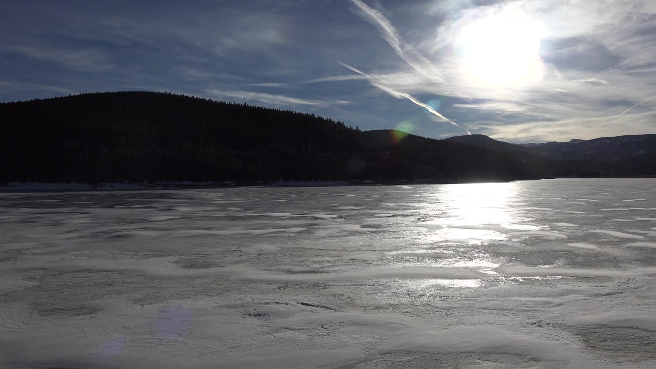 Bright sun breaks through thin clouds over a reflective foamy lake in Nederland, Colorado, with dark hills across the horizon and no visible people.