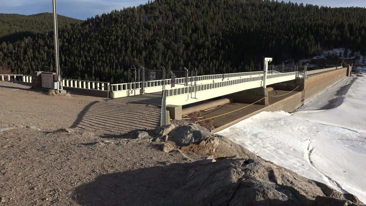 Opening frame of a Nederland, Colorado bridge over a frozen river channel, with gravel, foreground rocks, snow, evergreen-covered hills, and no visible people.