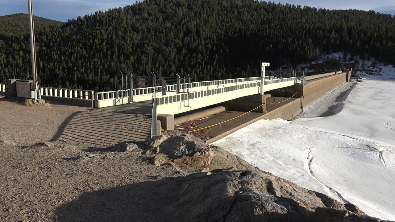 At 0:00, a white bridge crosses the edge of a frozen lake in Nederland, Colorado, with gravel, rocks, dark evergreen hills, and no visible people.