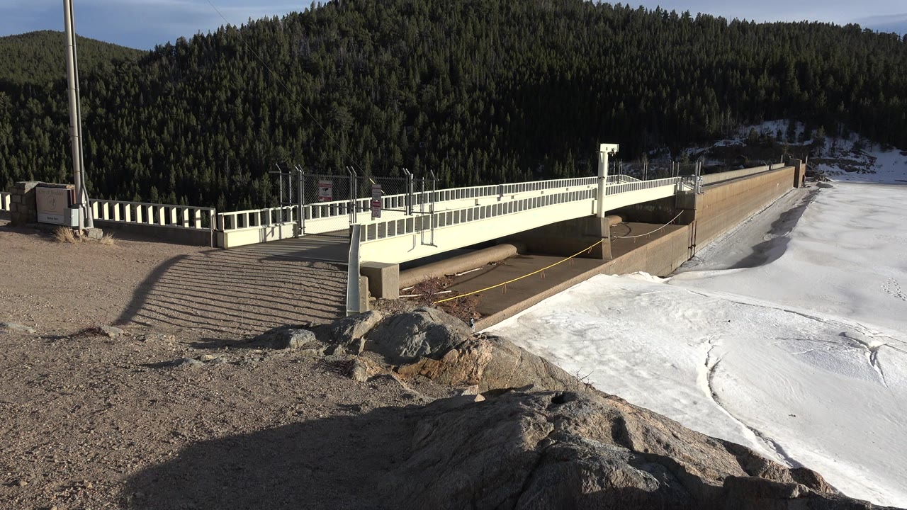 At 0:01, the white bridge and railings sit across the midground above the frozen lake in Nederland, Colorado, with rocks in front and no visible people.