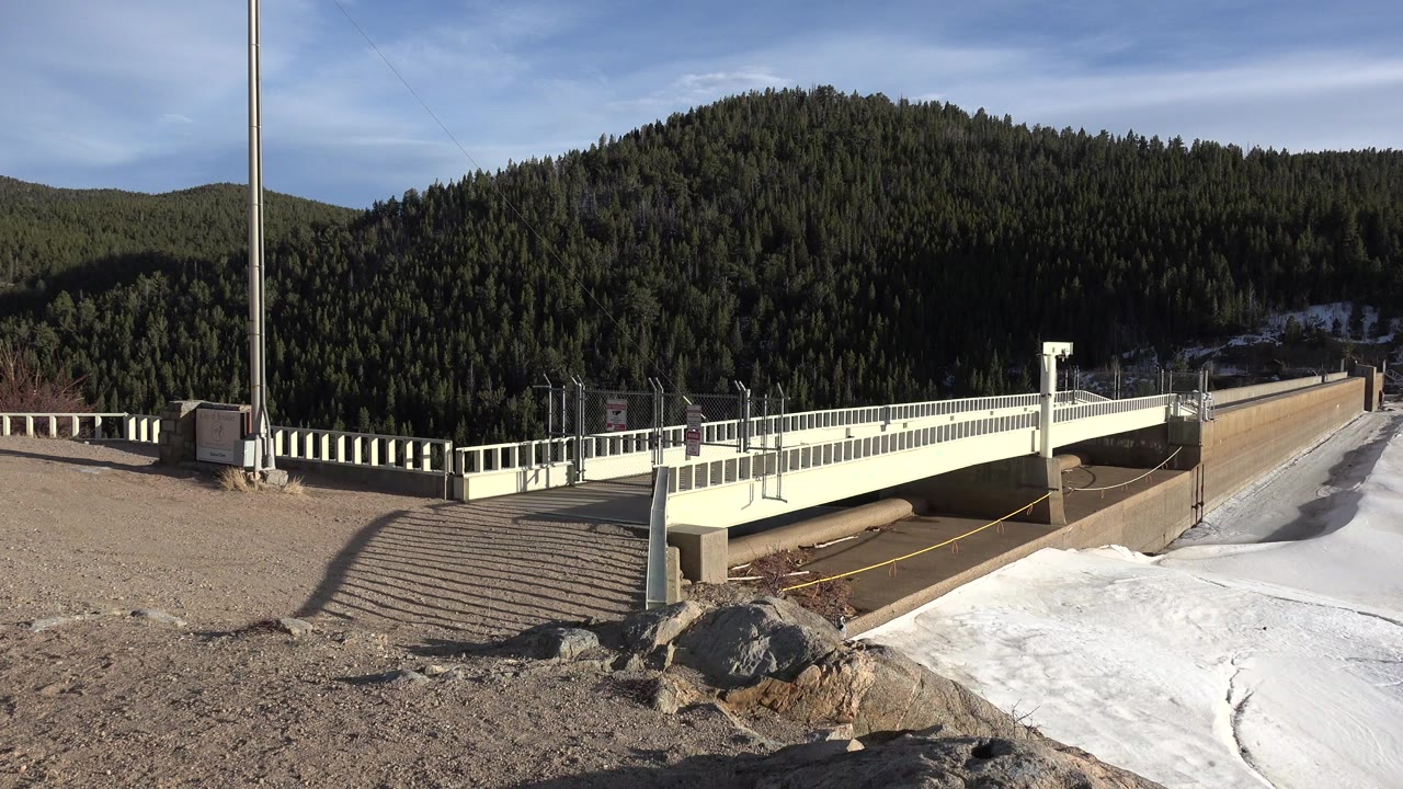 Wide Nederland, Colorado view of a pale bridge, gravel foreground, lingering snow patch, evergreen-covered slope, blue sky, and no visible people at the start of the clip.