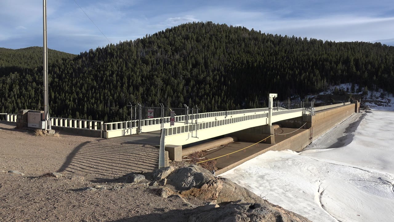 Second frame shows the bridge and concrete wall more centered beside the white snow or ice, with dark evergreen forest across the hillside and no visible people.