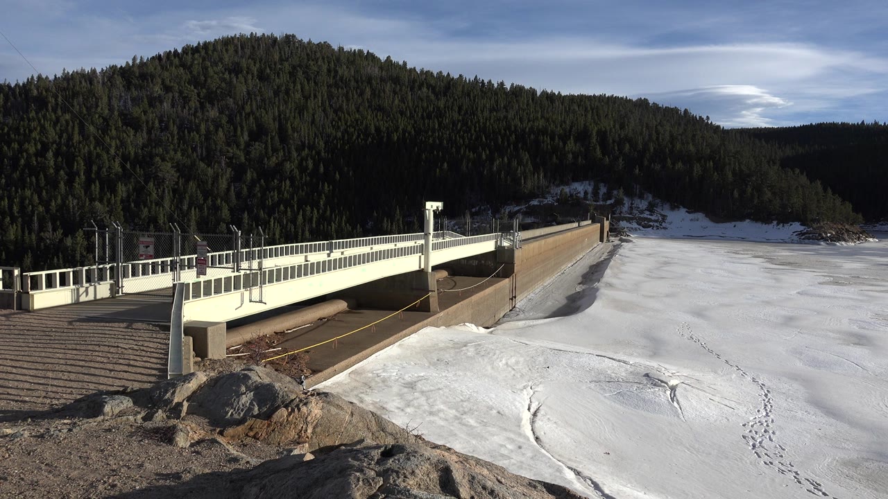Opening frame of a pale bridge over a snow-covered frozen basin in Nederland, Colorado, with rocks in the foreground, dark forested mountains behind, and no visible people.
