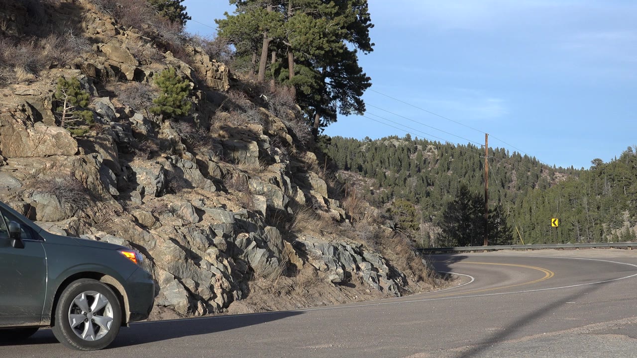 At 0:01 in Nederland, Colorado, more of the dark SUV crosses the lower frame while the road bends right past a rocky slope, guardrail, trees, and a yellow curve sign, no visible people.