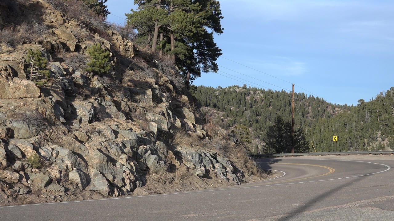 Opening frame of a paved road curving around a rocky slope in Nederland, Colorado, with pine-covered hills and no visible people.